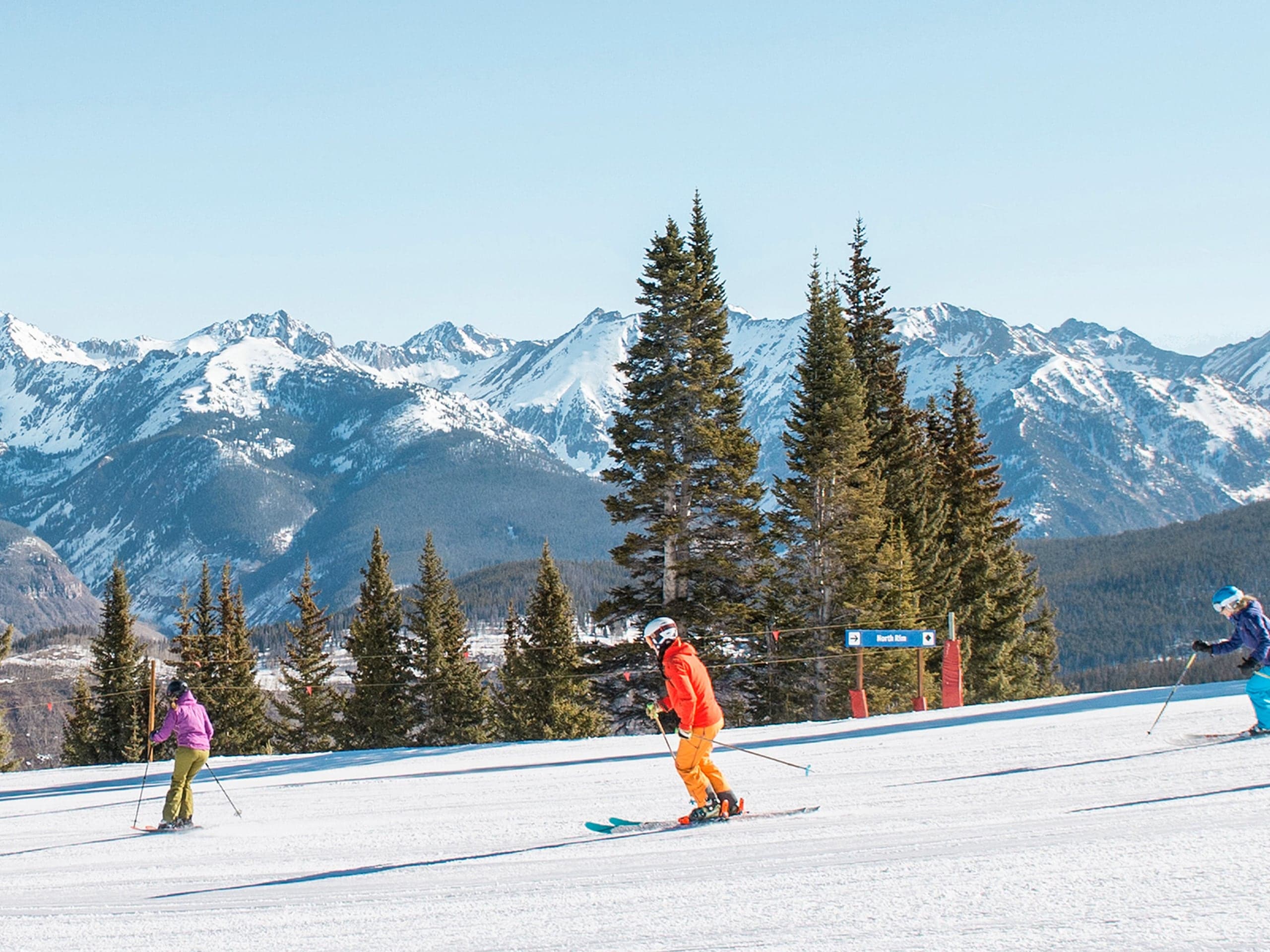Skiers on a groomed run beneath snowy Rocky Mountain peaks